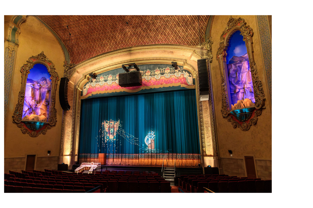 Interior of a beautifully decorated theater with an ornate stage, blue curtain, and a ceiling adorned with intricate patterns. Seating area consists of red chairs.