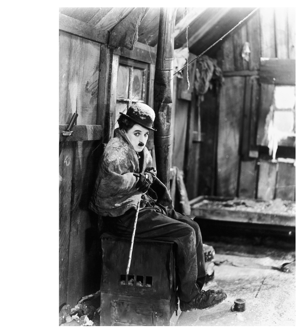 Charlie Chaplin, in character with a solemn expression, sits on a crate in a rustic wooden shack, holding a cane and wearing a bowler hat and tattered clothes.