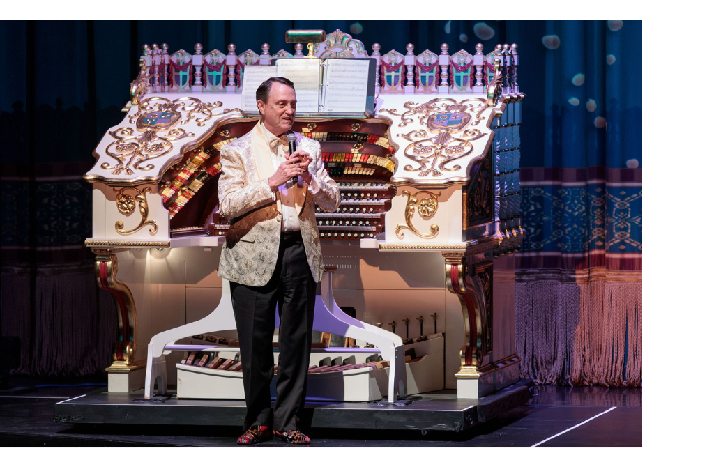 A person stands center stage holding a microphone, with a large, ornate theatre organ in the background. The individual is dressed in a formal, patterned jacket and black pants, addressing the audience at a live performance venue.