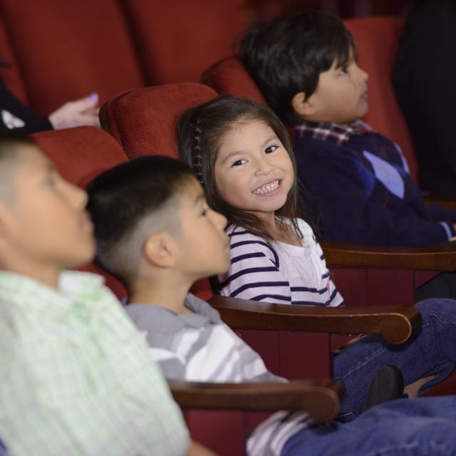 A young girl, seated in a row with other students at a theatre, smiles at the camera.